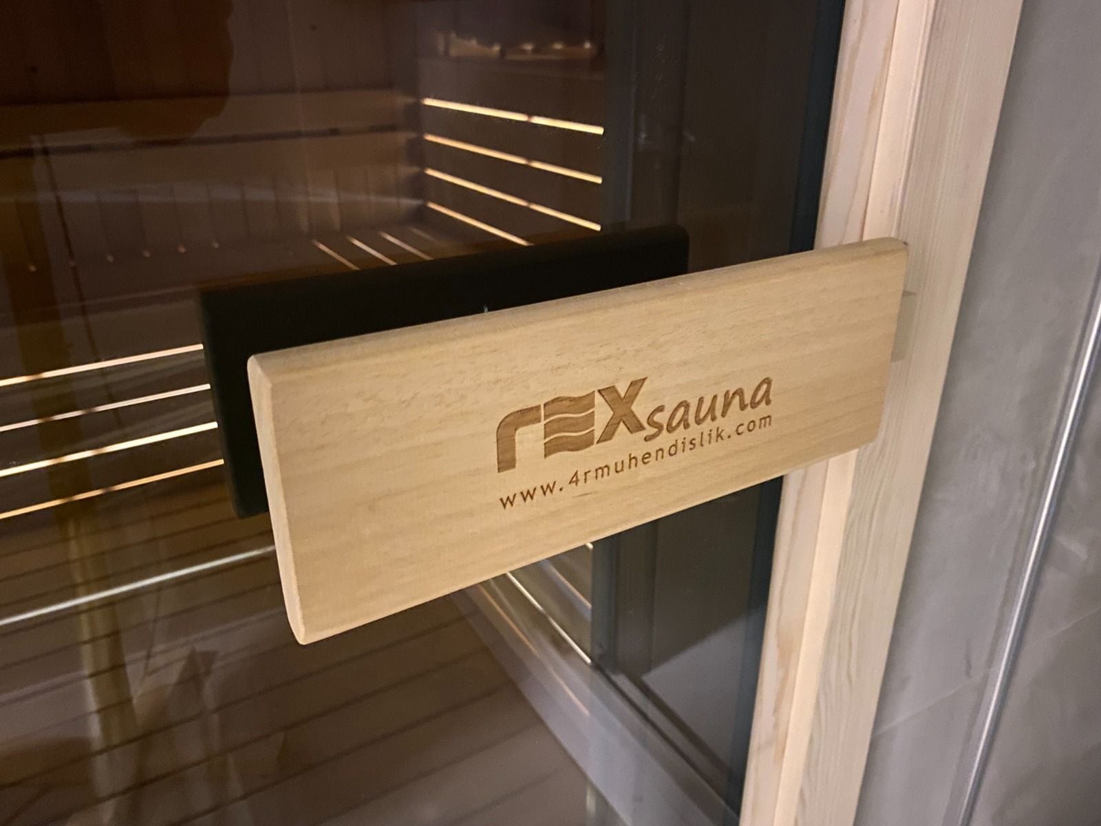 rexsauna logolu ahşap kapı kolu, cam kapının ardında aydınlatmalı sıralar